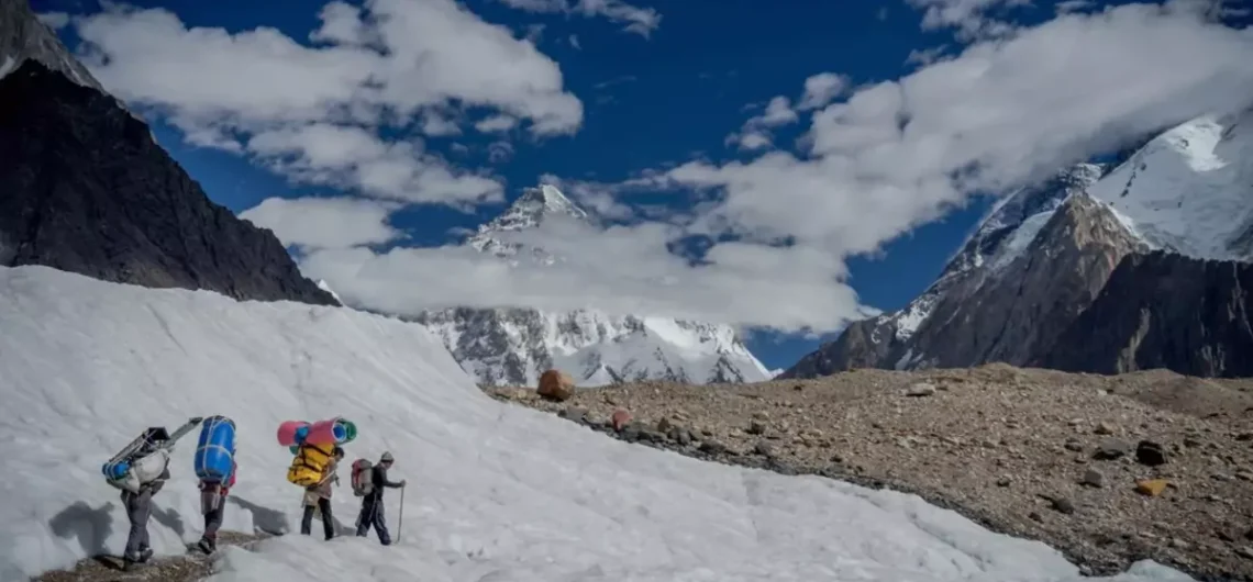 Trekkers and porters with colorful gear ascending a snowy glacier path towards K2 Base Camp, with K2 visible in the distant Karakoram mountains.