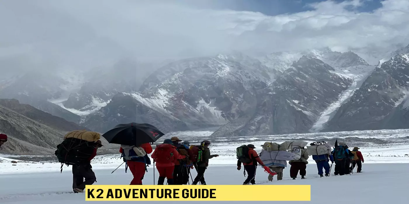 pakistan-k2-expedition-journey A scenic shot of a group of adventurers and their support team making their way across a snowfield with massive mountains behind them